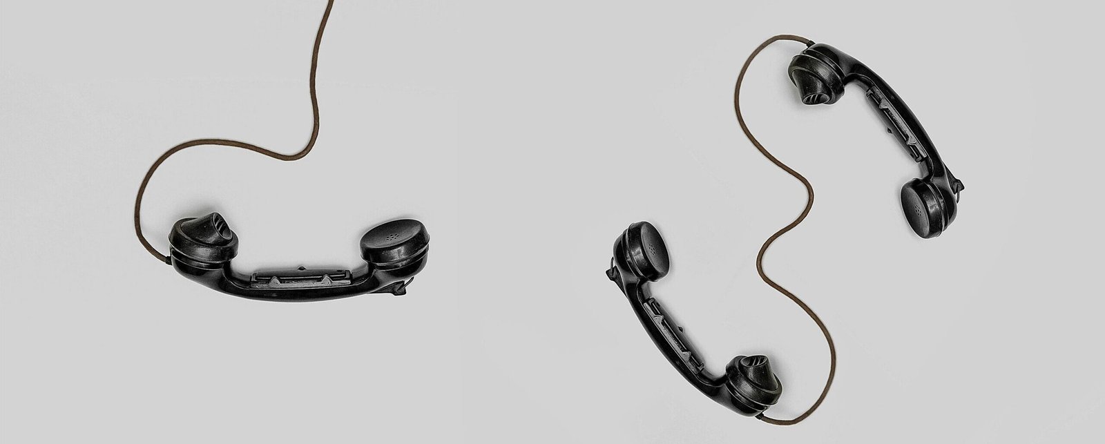 Two vintage black telephone handsets connected by cords on gray background.